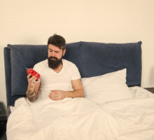 Bearded man in bed with red alarm clock is ready to go to sleep